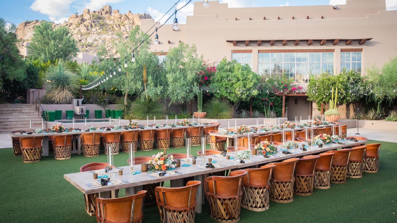 Three long tables outside with dark orange chairs on green grass next to a beige building.