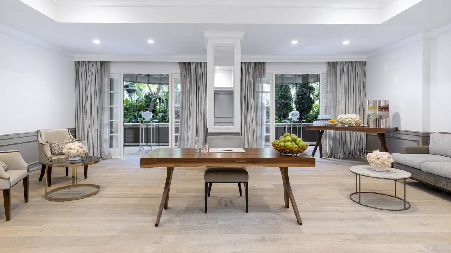 A seating room with a desk, two chairs, a sofa, a table with refreshments and large windows.