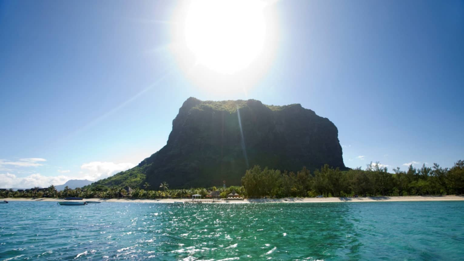 Long view of a forested monolith mountain sitting atop crystal waters. Sun beams down from above in a clear blue sky.