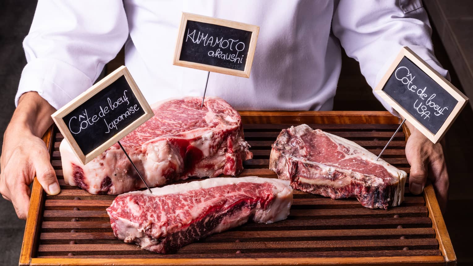 Close-up view of a wood tray with three labelled cuts of well-marbled beef, held by person in white chef's coat.