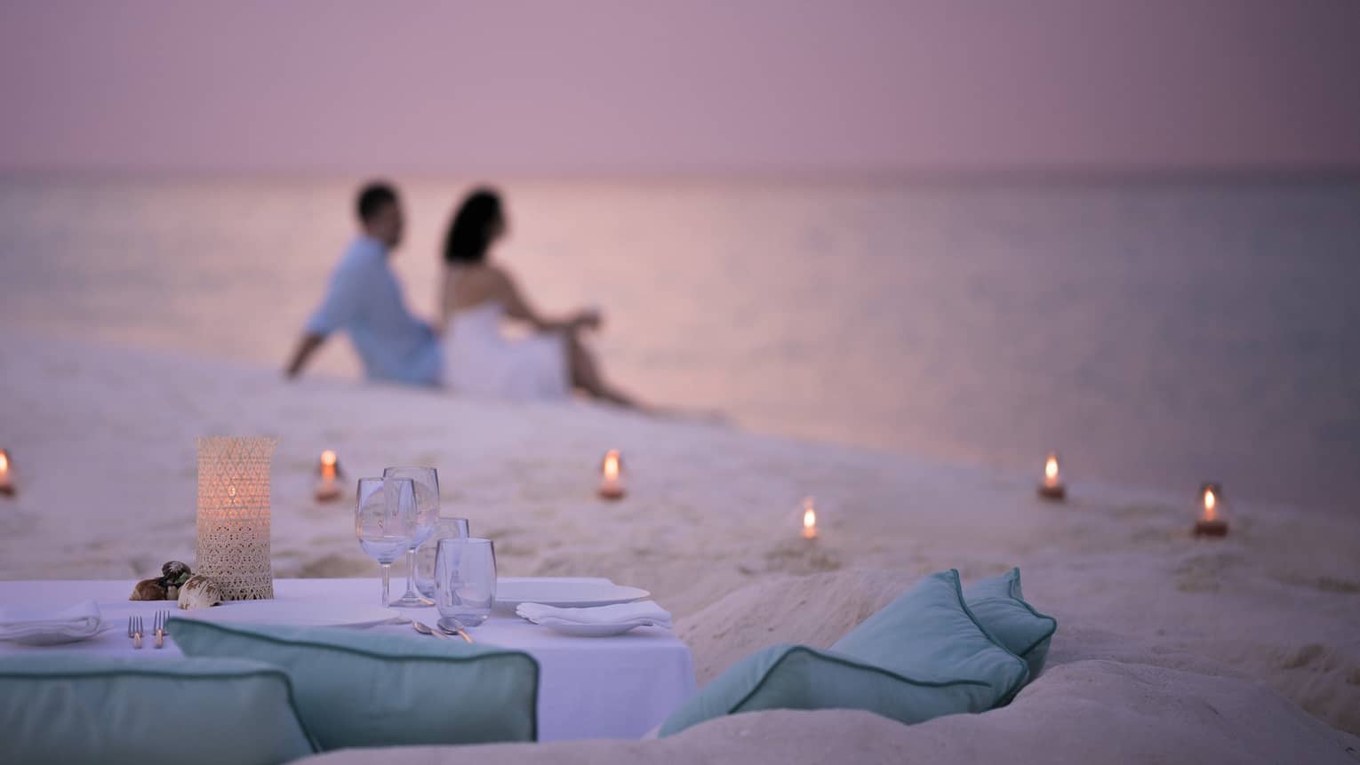 Private dining table in front of sand beach with tealight candles, couple sitting in sand