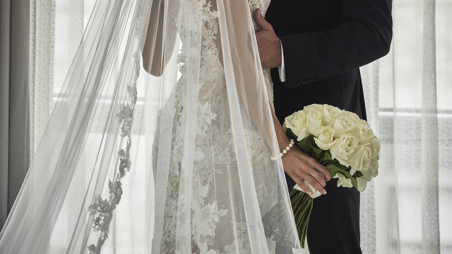 Groom and bride with long lace veil and white wedding bouquet embrace