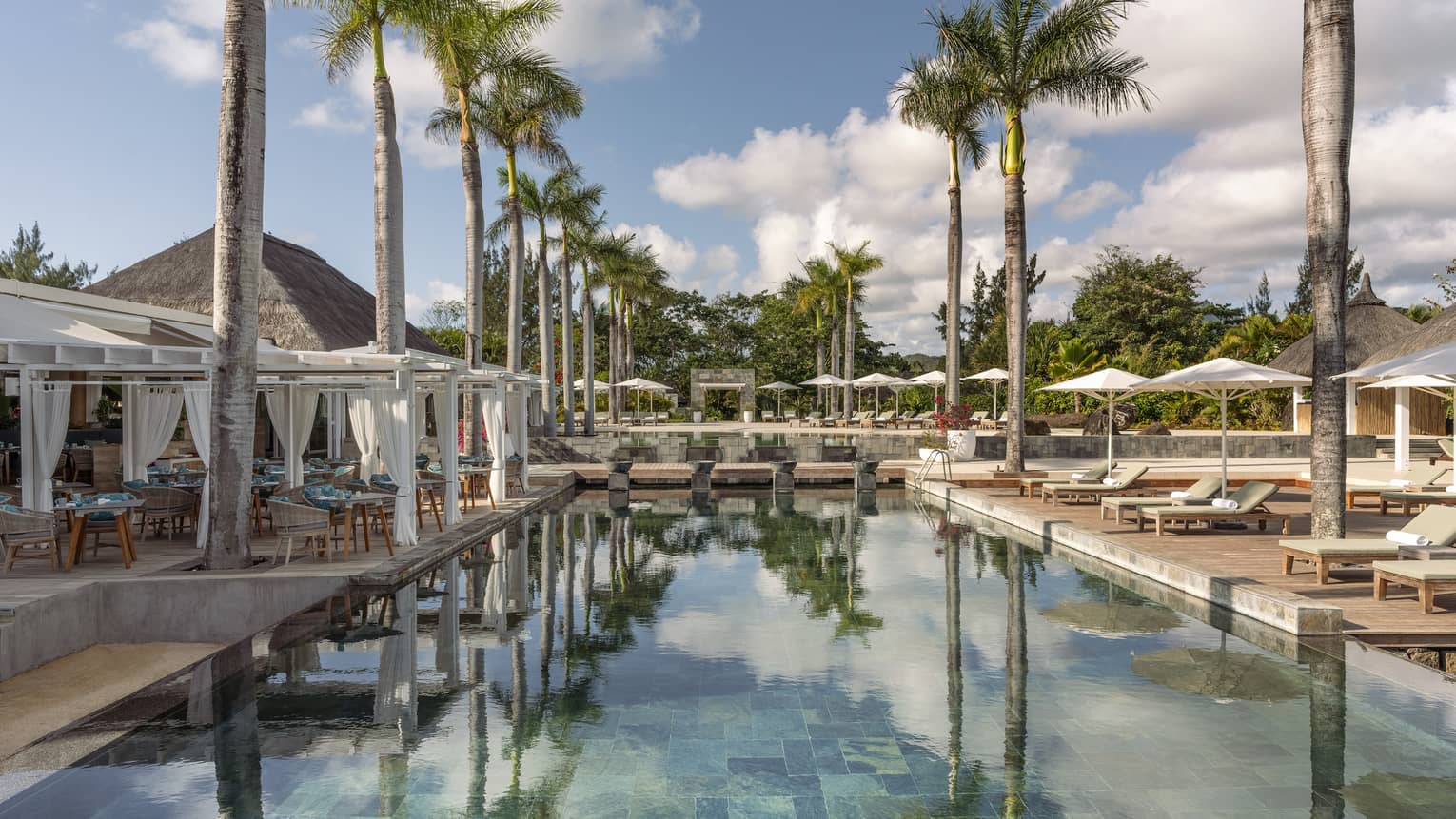 Outdoor pool flanked by palm trees, cabanas and lounge chairs with umbrellas