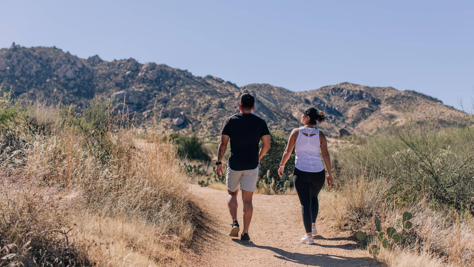 A man and woman in casual clothing walking along brush towards hills in the distance.