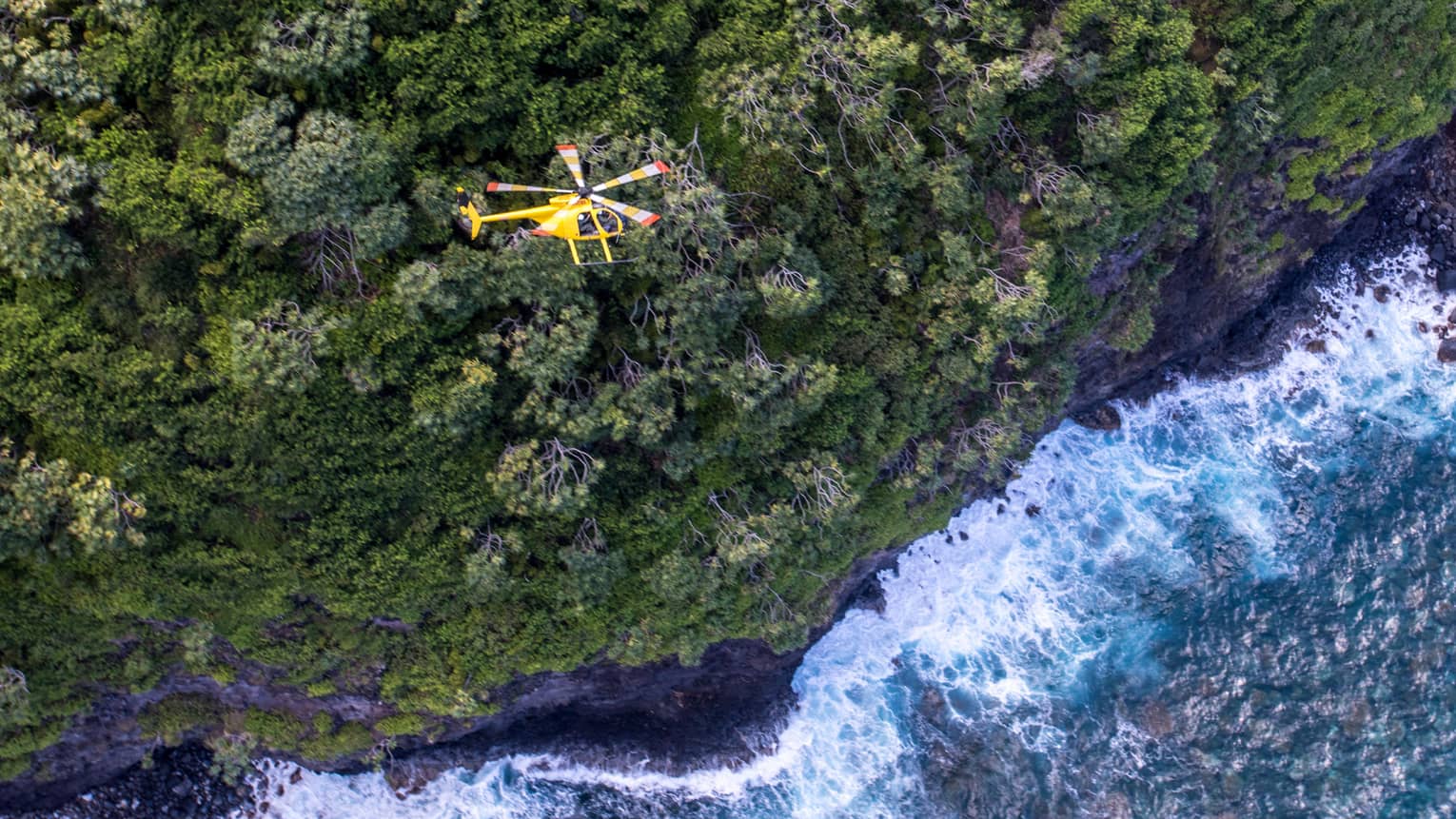 A yellow helicopter flies over a steep forested mountain. Powerful waves crash against the shoreline in the water below.