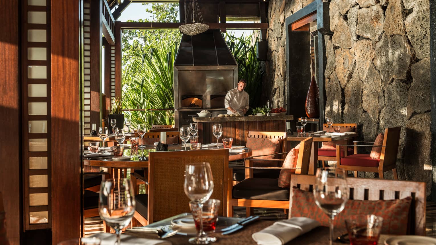 Chef works at counter in front of steel oven at end of bright dining room, table with wine glasses