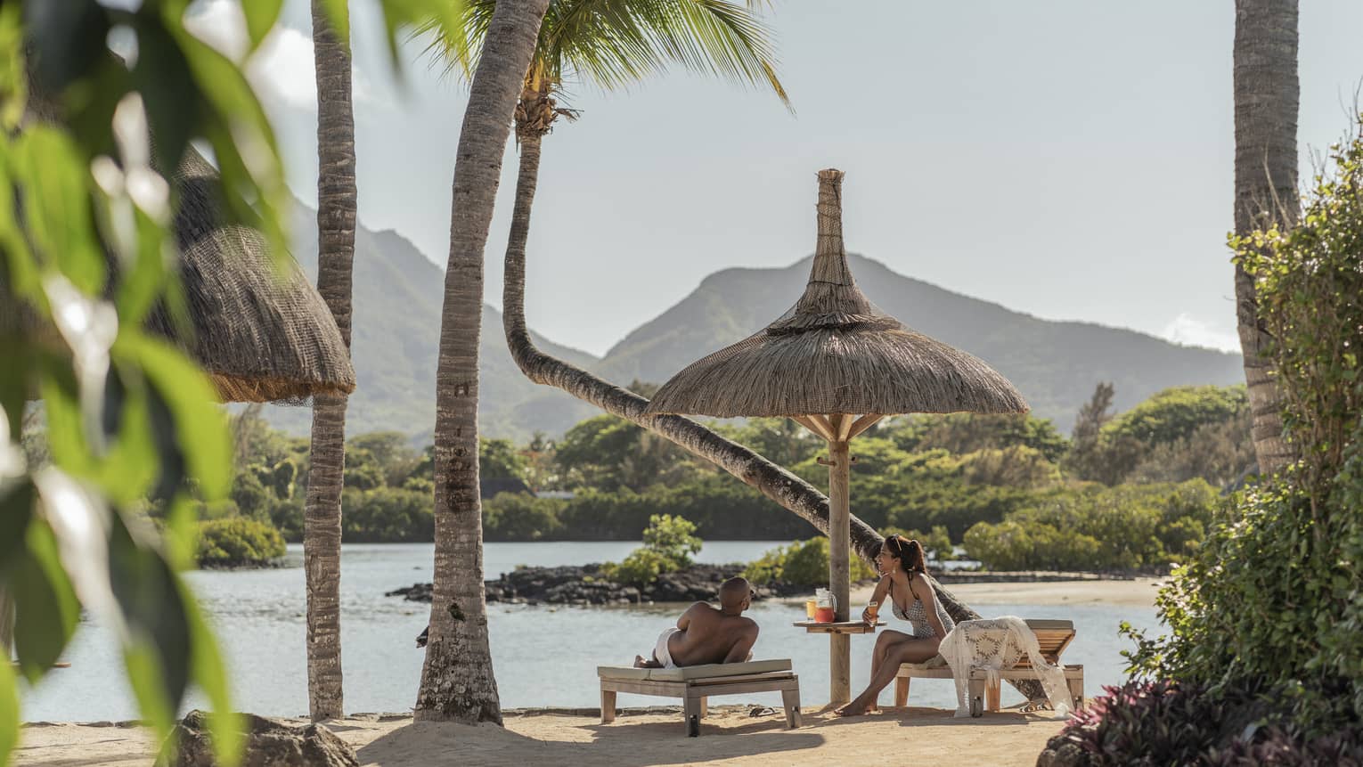 Couple relaxes on oceanfront chaise lounges, mountains and palm trees in the background