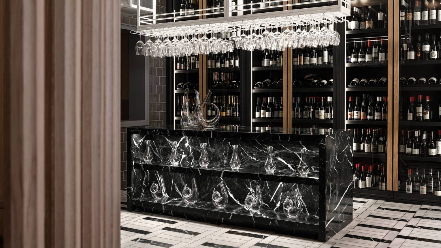 A wall lined from floor-to-ceiling with wine bottles sits behind a black marble counter that holds two shelves of glass decanters with hanging stemmed wine glasses hanging overhead