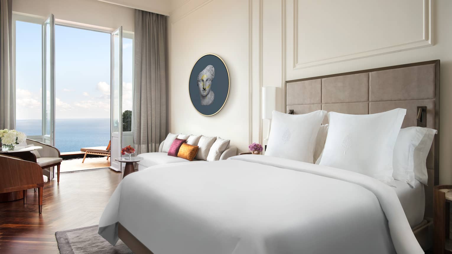 Hotel room with white king bed, white walls with crown molding, wood floors, walk-out sea-view balcony