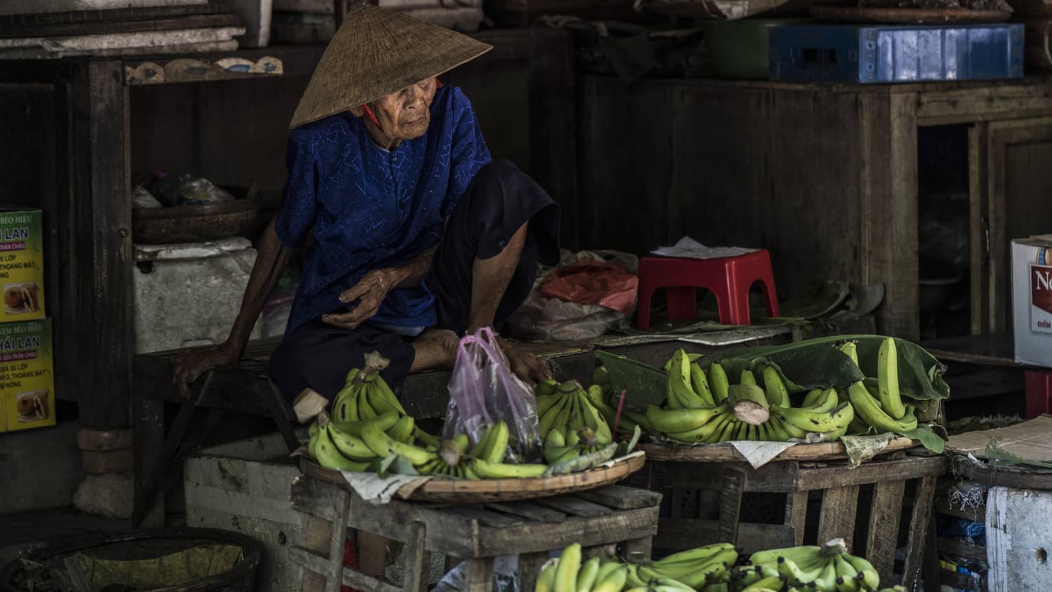 An elderly farmer wearing a blue outfit and woven straw hat selling bananas at a farm stand in Hoi An Historical Town