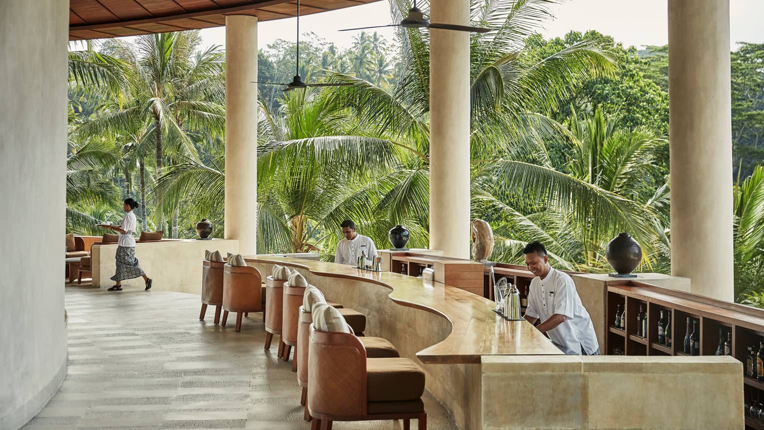 Smiling bartenders behind long wood-and-stone bar under white pillars of resort balcony, palm trees in background