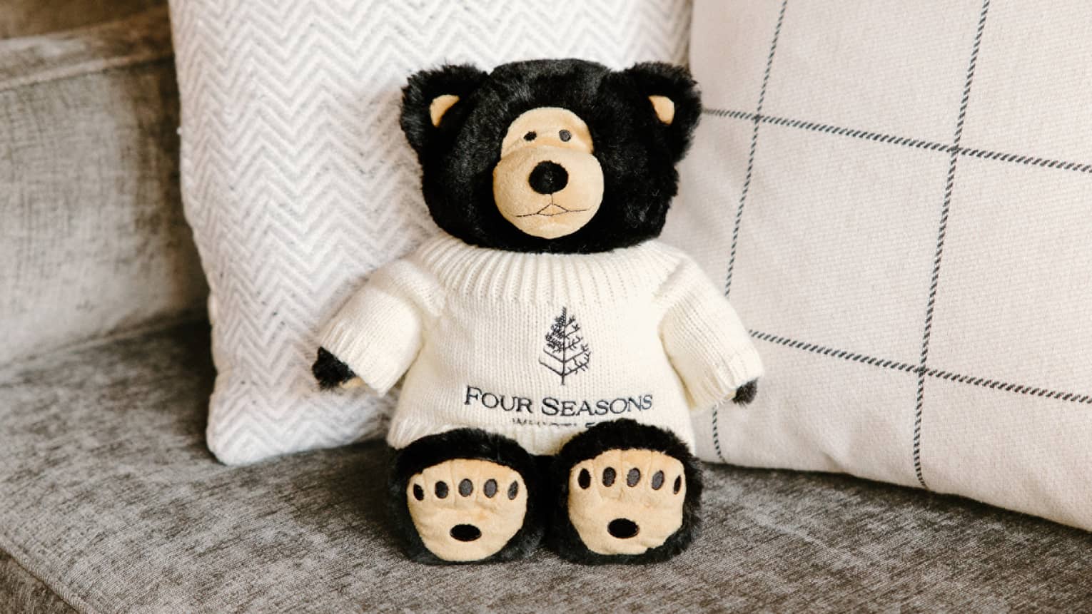 Stuffed black bear in cream-coloured sweater propped up on grey couch