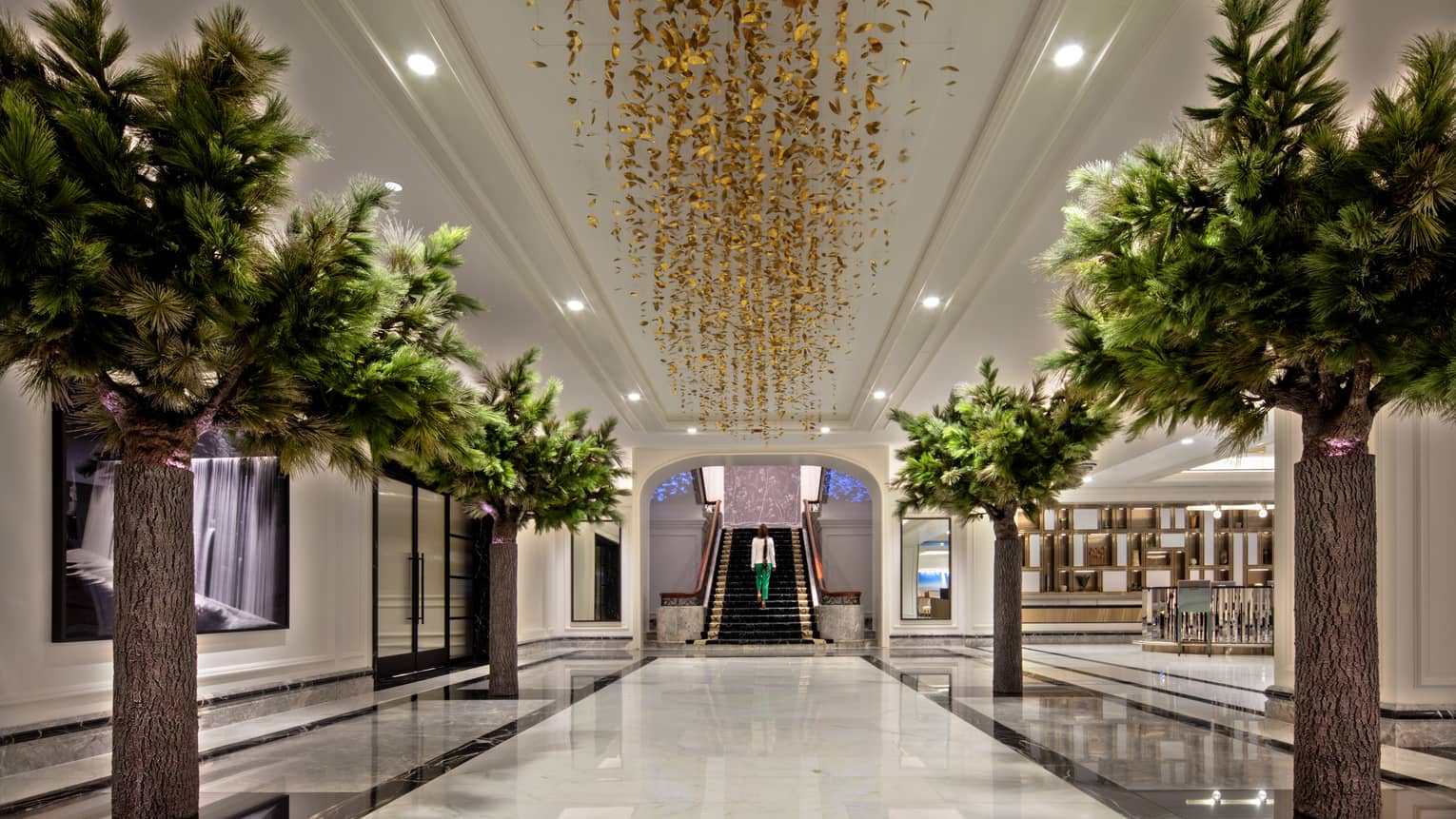 The hall of a hotel property with a gold leaf chandelier, green plants lining the hall and stairs leading up at the end.