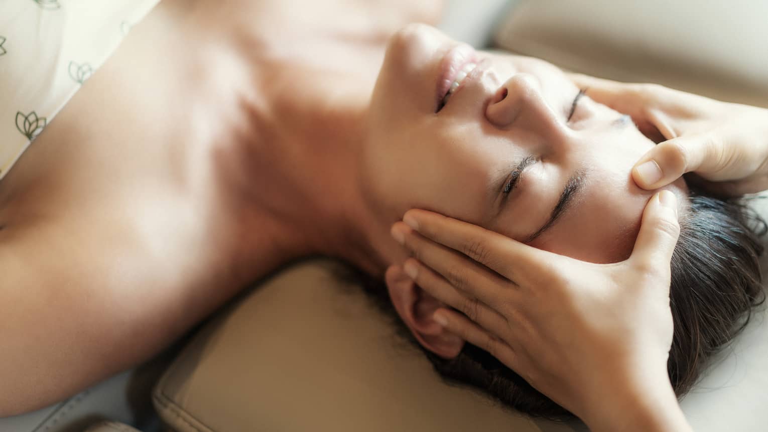 Spa facial, two hands rest on woman's forehead as she closes her eyes, lays under sheet on treatment table