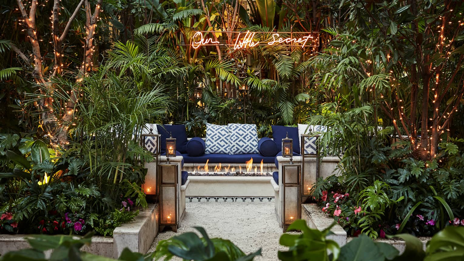 Our Little Secret sign over cushioned bench behind outdoor fireplace, lanterns, tropical garden