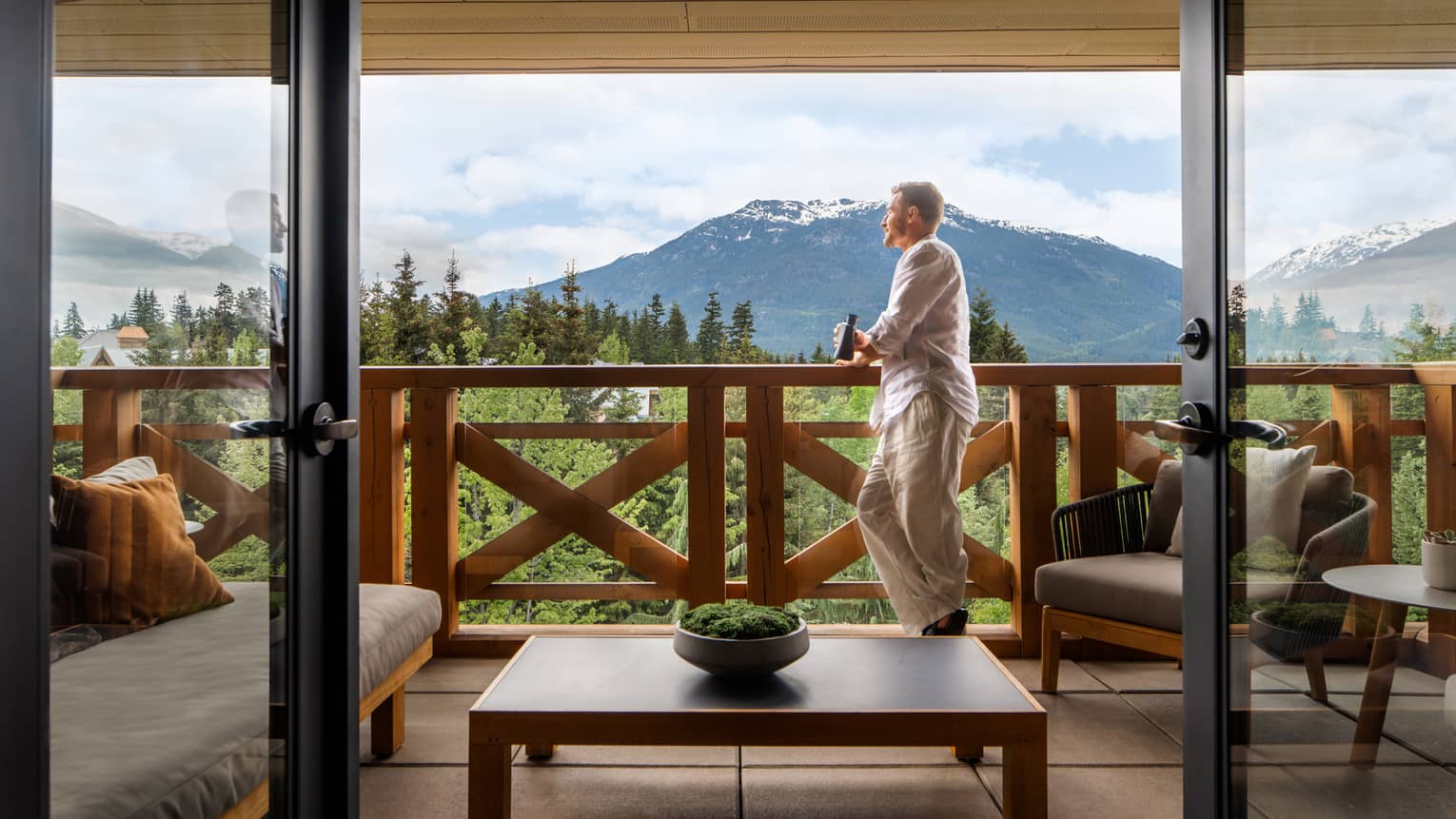 Man with short hair wearing a white long-sleeve shirt and khaki pants leans on the railing of a balcony with a beer in hand, looking out onto the mountain views