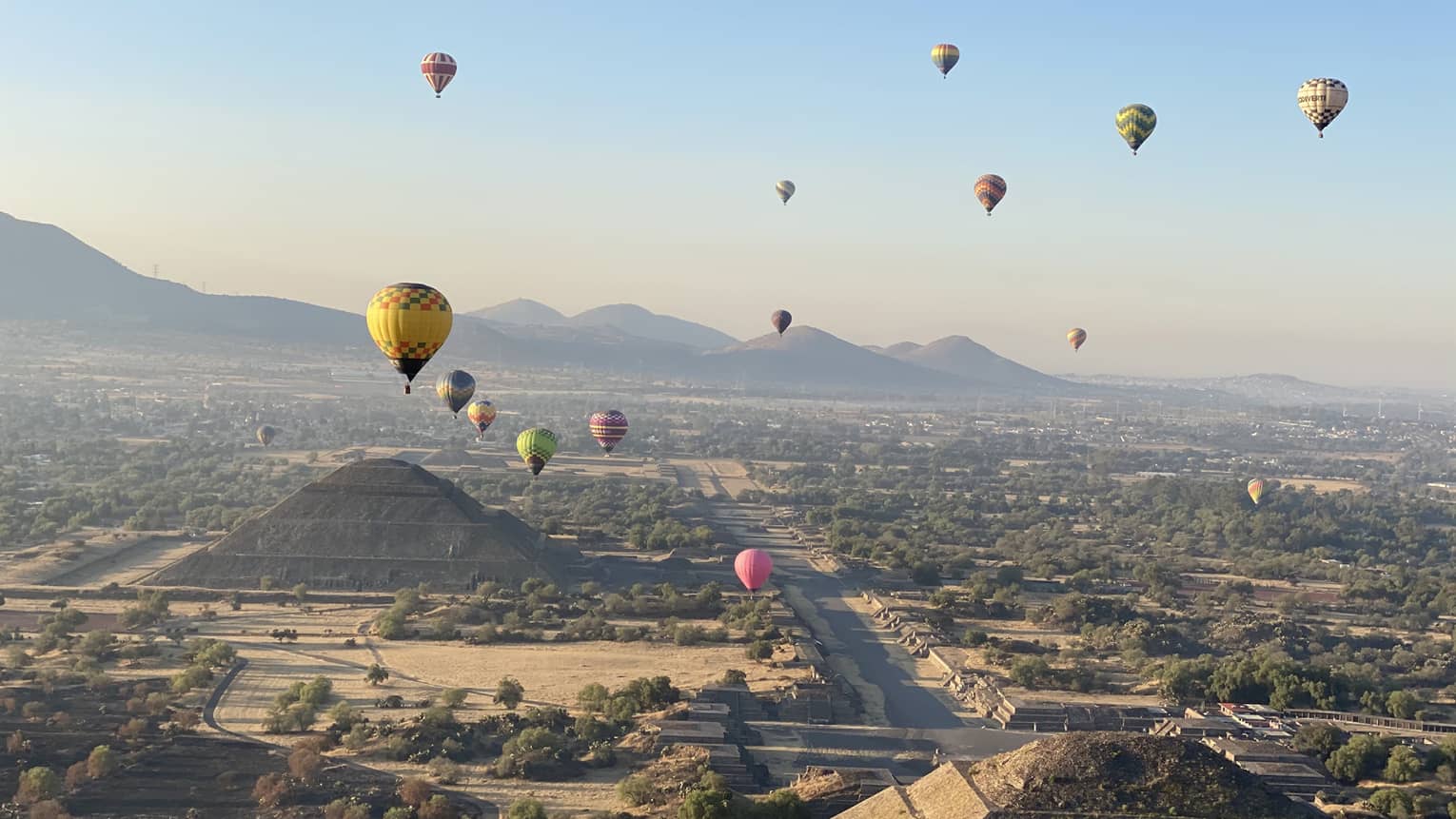 Colourful hot air balloons float above the Teotihuacan pyramids with hills in the distance on a clear, sunny day
