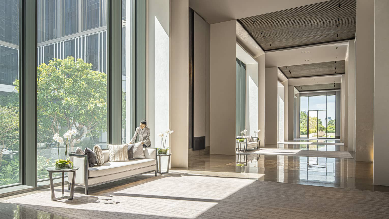 Staff member fluffs the couch pillows in the Ballroom Foyer with cream-coloured carpeting and furniture, floor-to-ceiling windows