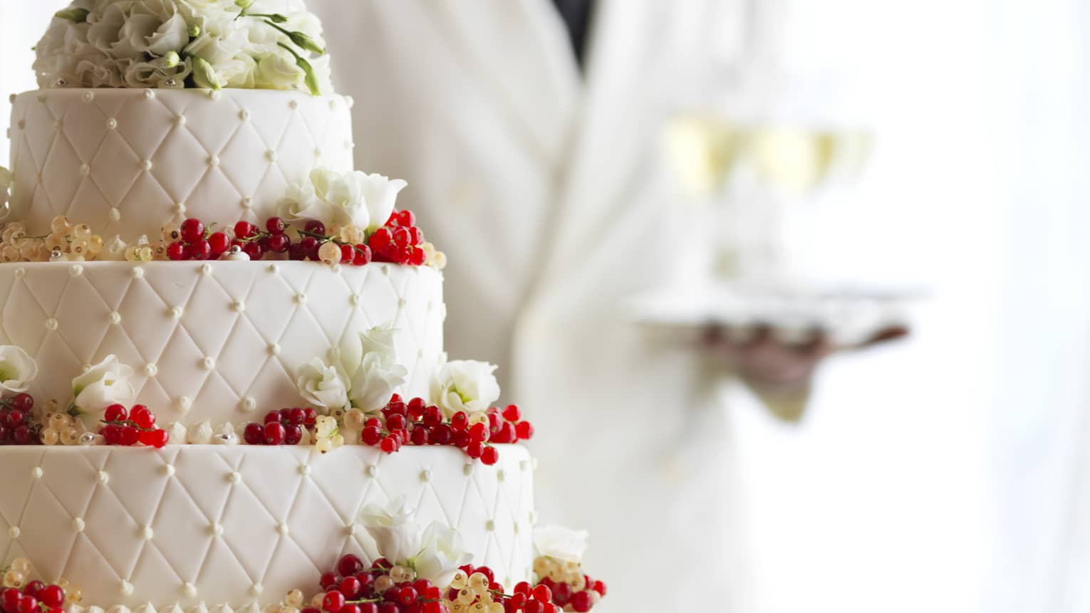 Waiter with tray of Champagne glasses behind white tiered wedding cake with flowers