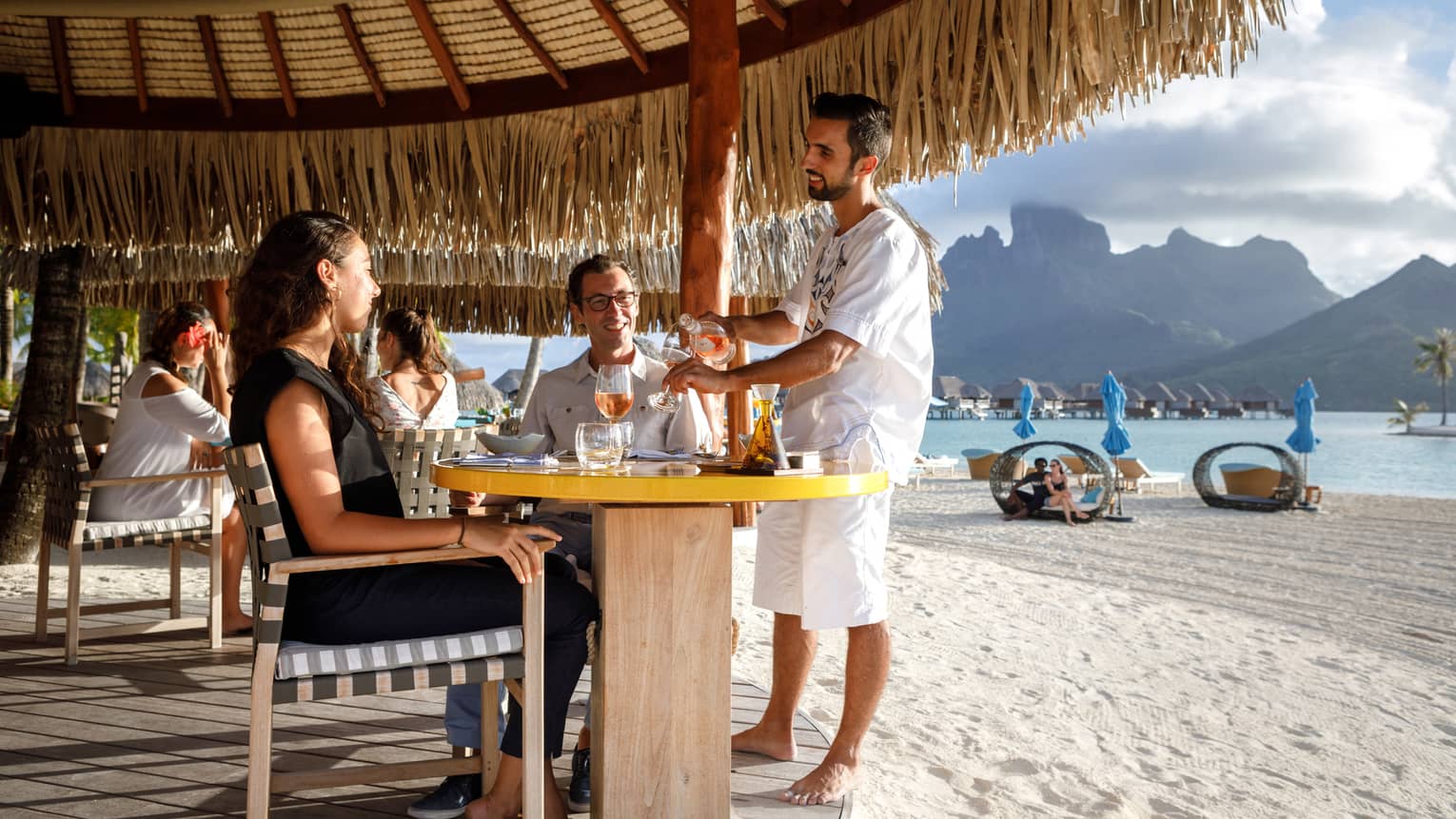 Staff refills wine for couple at patio table on white sand beach