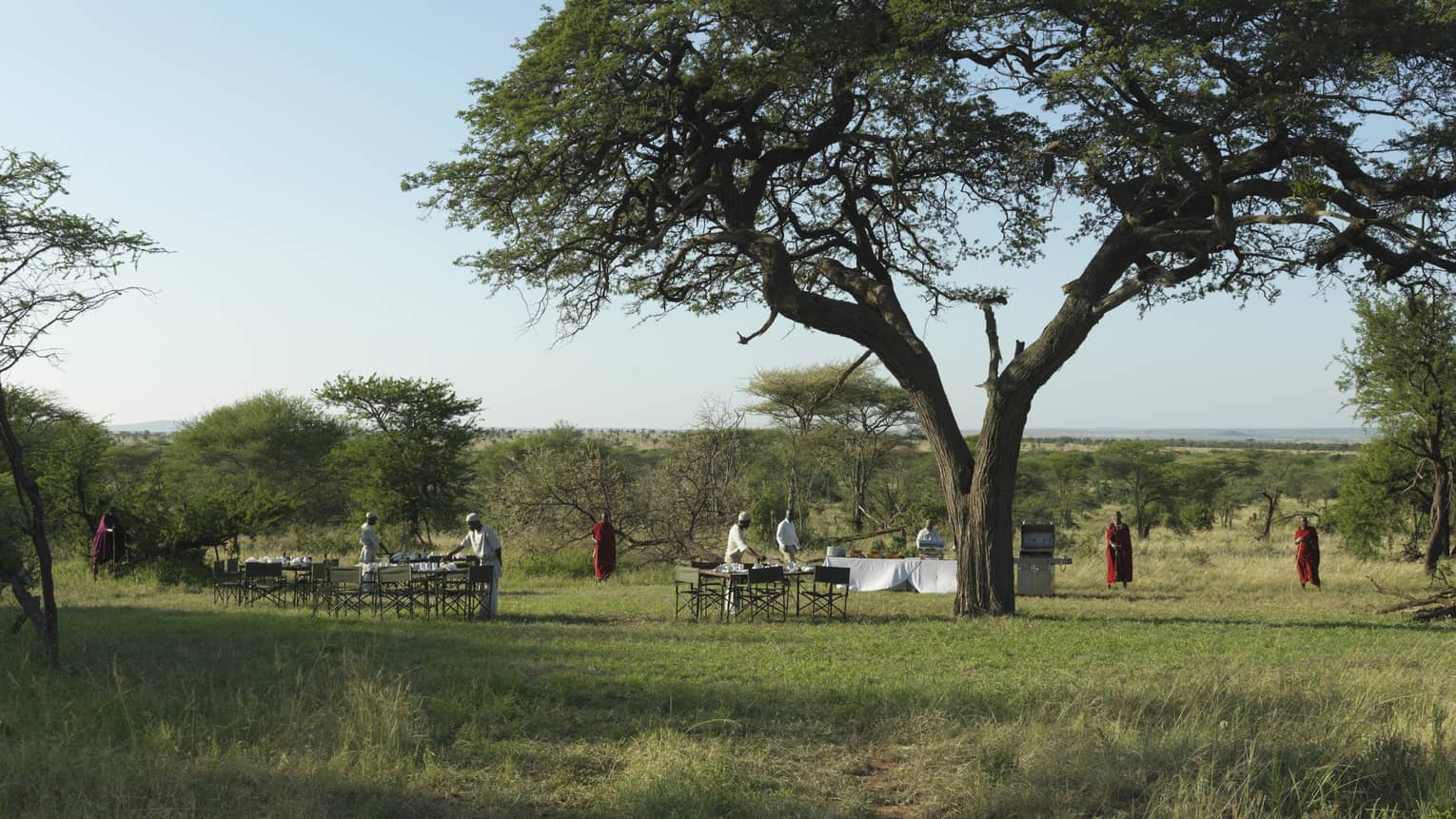 Staff set outdoor table and chairs as Maasai stand nearby at the outdoor meeting pit
