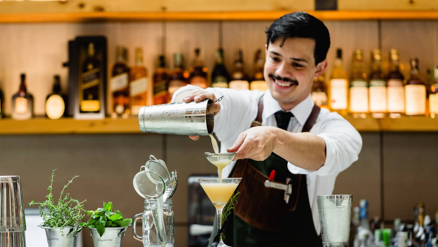 Smiling bartender pours orange cocktail from shaker into martini glass at bar 