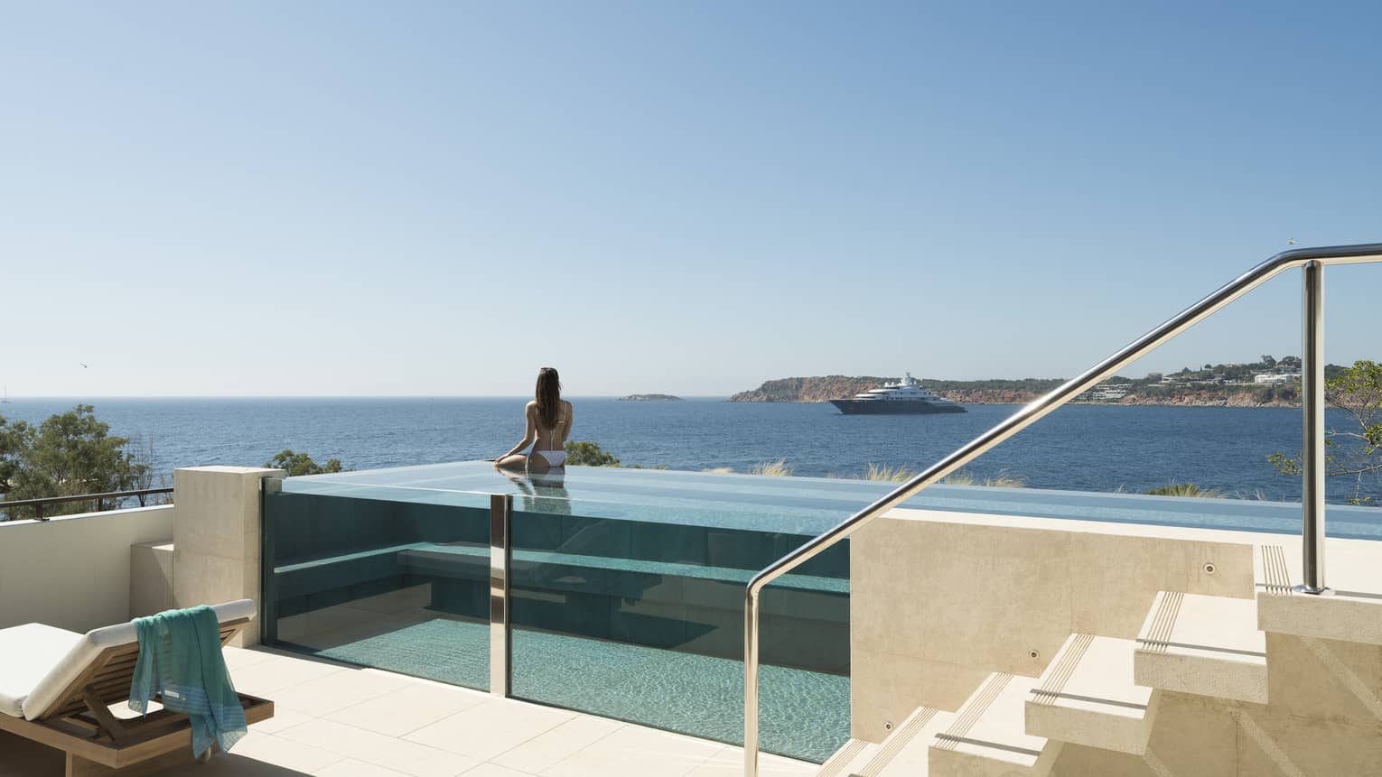 Terrace with staircase and glass infinity pool, a woman wearing white bikini in pool looks out to sea