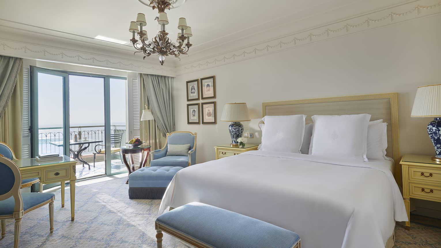 Bedroom with white king bed, chandelier, light blue bench and arm chair, and a balcony overlooking the sea