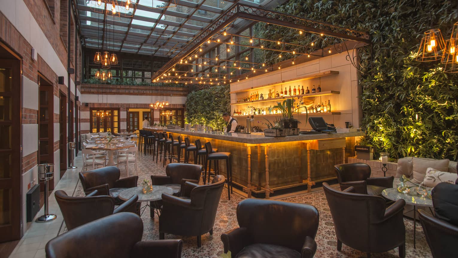 A bar counter surrounded by twinkle lights is set in front of a green wall filled made up of live plants, with numerous round cocktail tables and brown leather club chairs in the foreground 