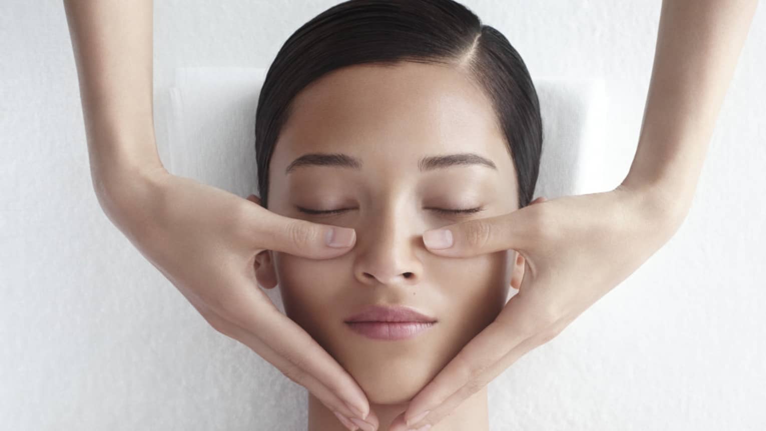 Woman in white bathrobe lies on massage table with eyes closed, masseuse rests hands on her cheeks