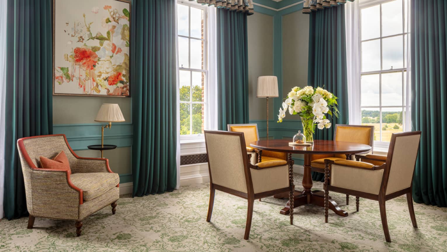 Guest room with round table and four dining chairs, tan accent chair, twin windows with blue drapes