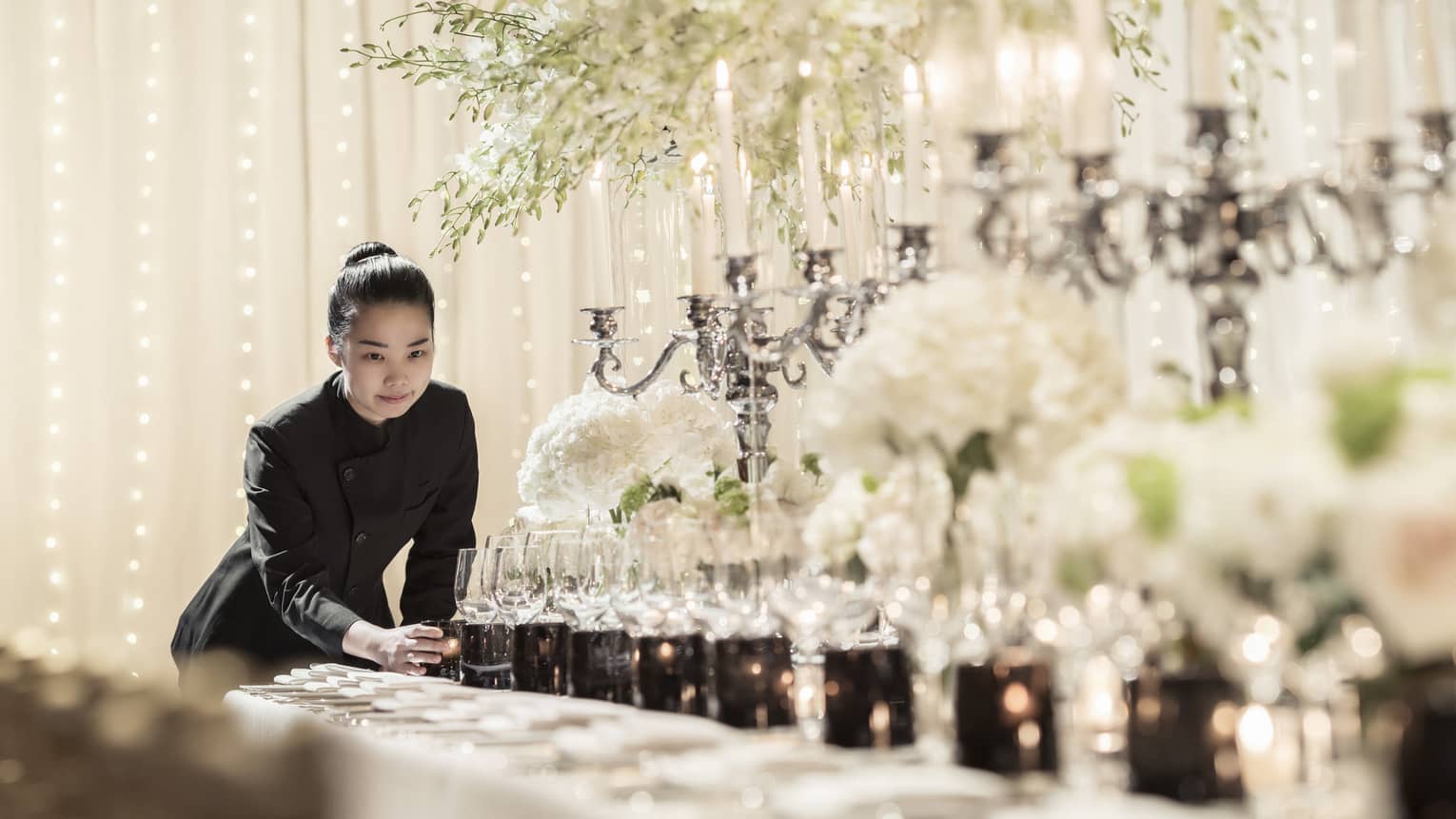Hotel staff places candle at end of long formal dining table set with candelabras, sparkling lights