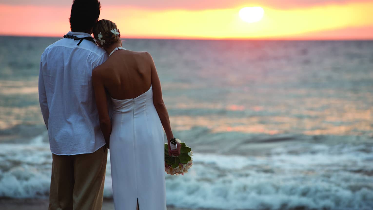 Back of groom and bride in white dress with bouquet look out over beach, sunset