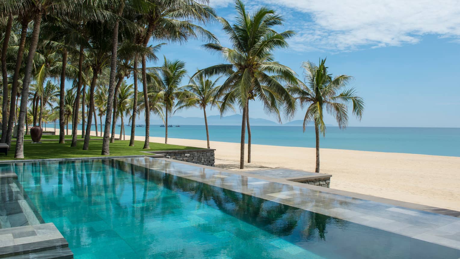 Tall palm trees between stone outdoor swimming pool and white sand beach