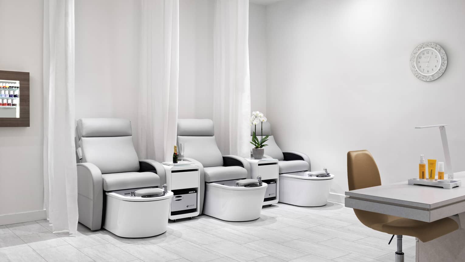 Nail salon with three light grey pedicure chairs, separate by white curtains, and one manicure station