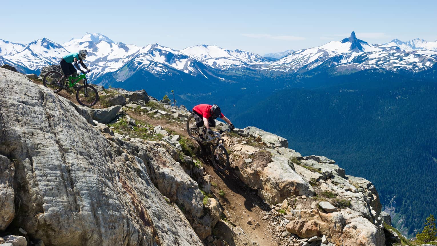 Two people on mountain bikes cycle down rocky mountain near peak, snow caps in background