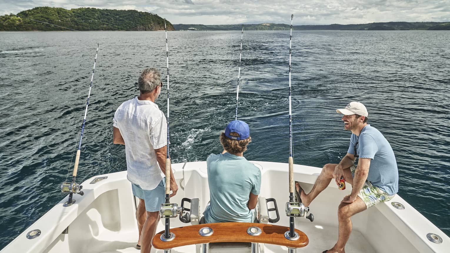 Three men of varying ages sit looking out at the water from the back of a boat outfitted with fishing rods