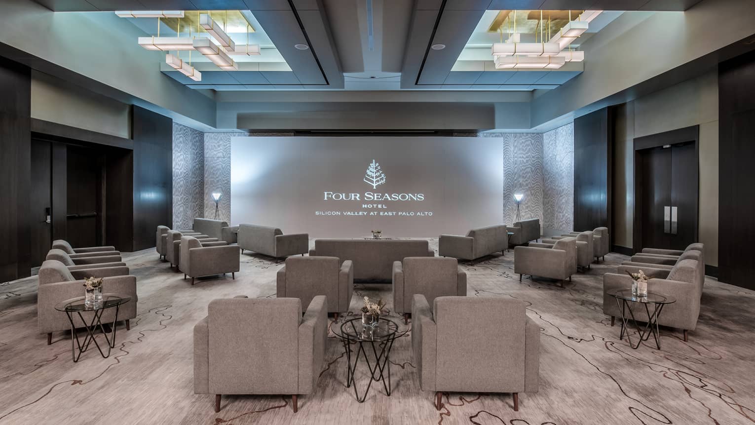 Gray plush chairs are angled towards a large projector in an indoor carpeted event space