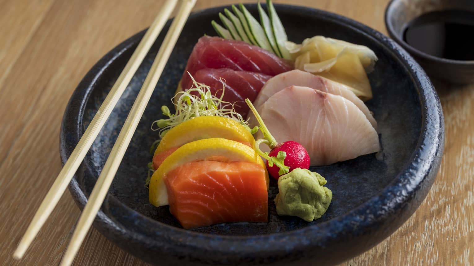 Tuna, salmon and hamachi sashimi served in a black stoneware bowl with chopsticks on the side