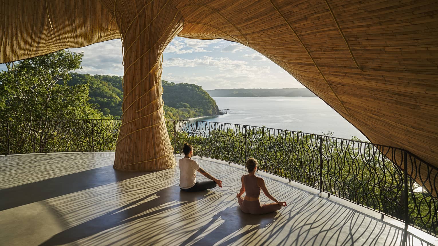 Two people sit with legs crossed and hands on knees on the floor of an open-air wellness studio that overlooks the ocean