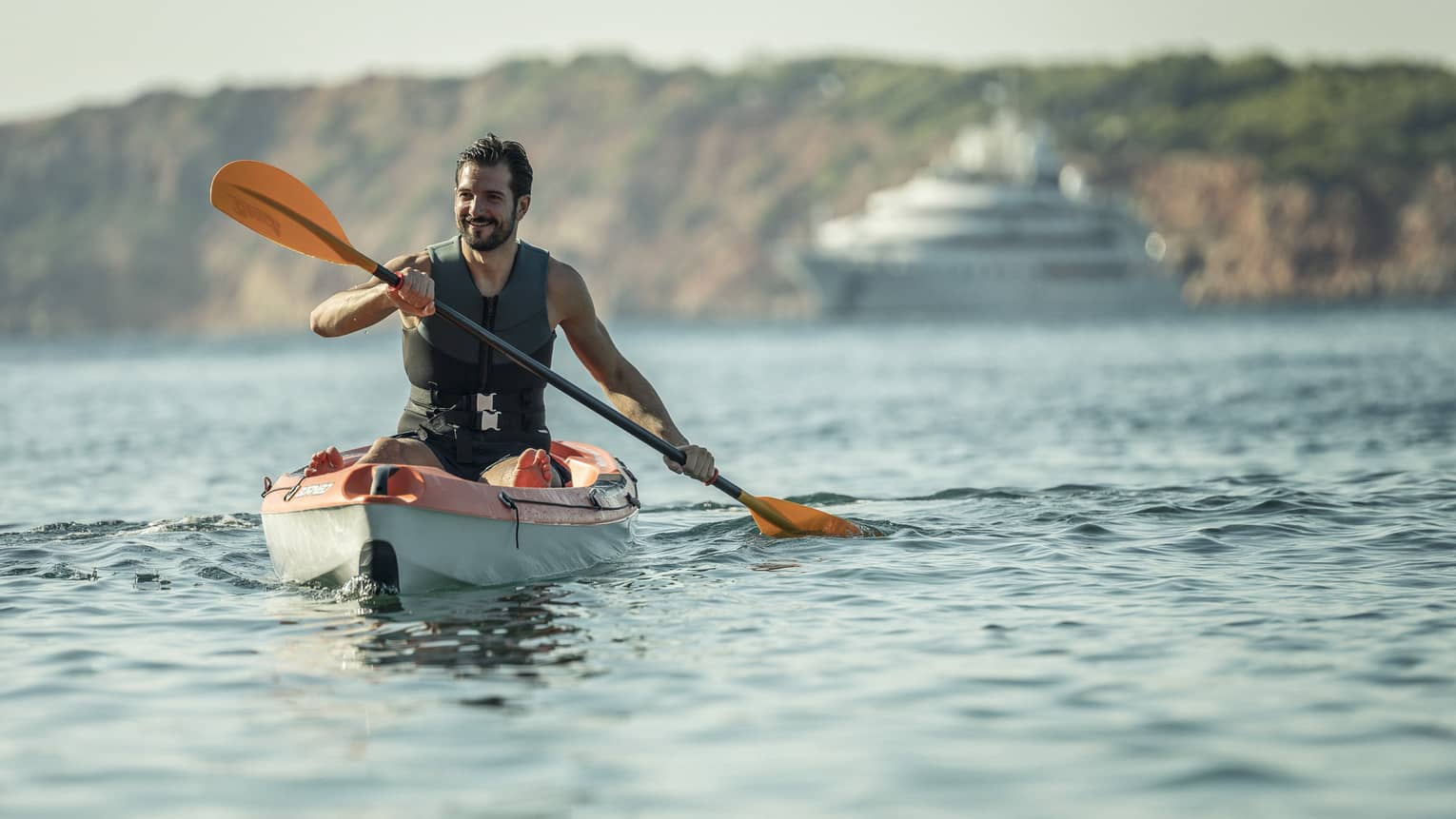 Man smiling while kayaking in sea, yacht and rocky coast in backdrop