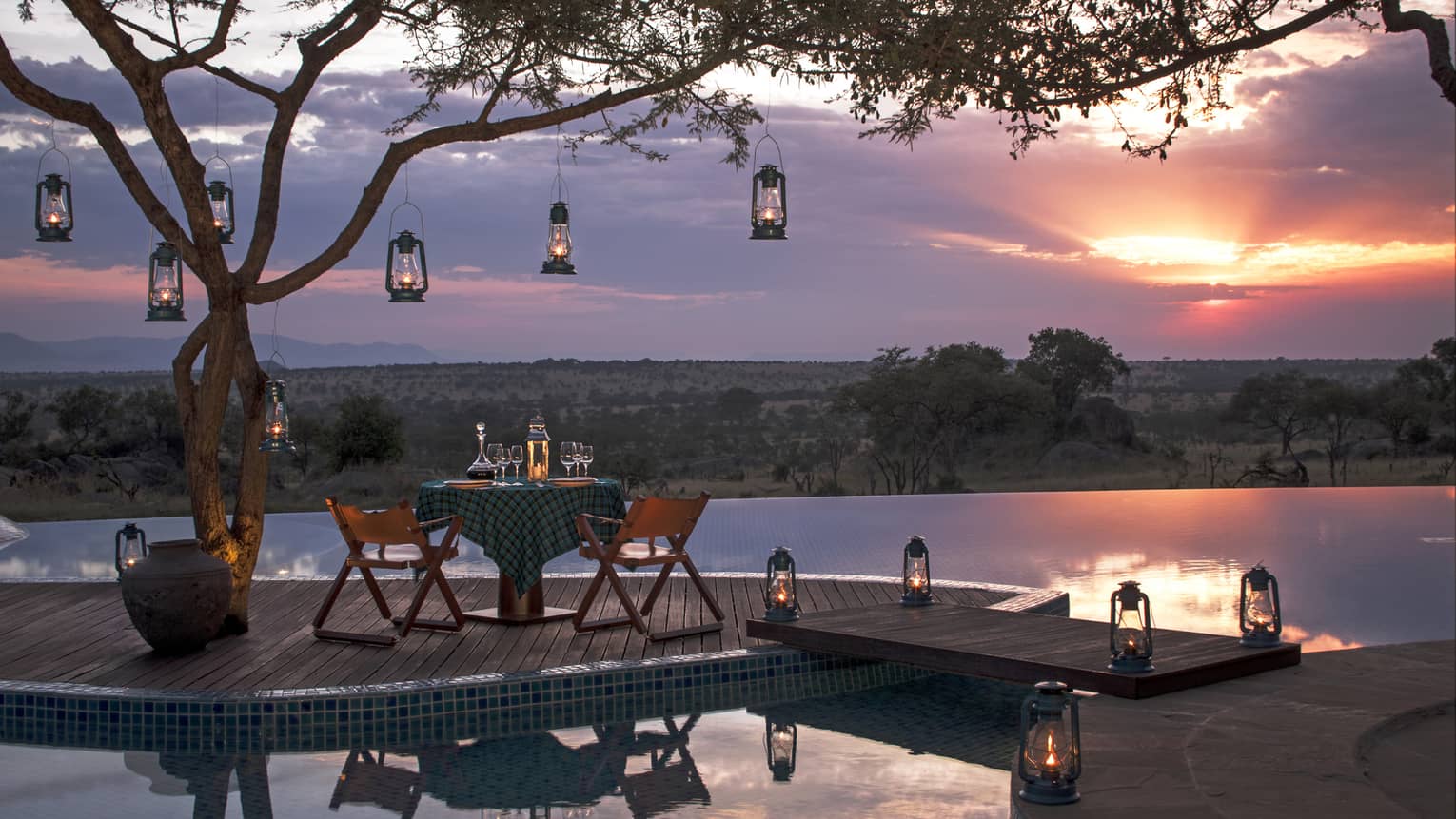 Private dining table on wood dock lined with lanterns, hanging from tree, sunset