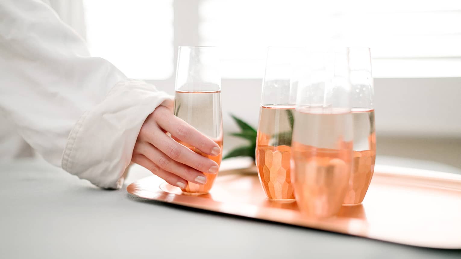 Woman in white bathrobe hand reaches for glass of water, next to three glasses on rose gold tray