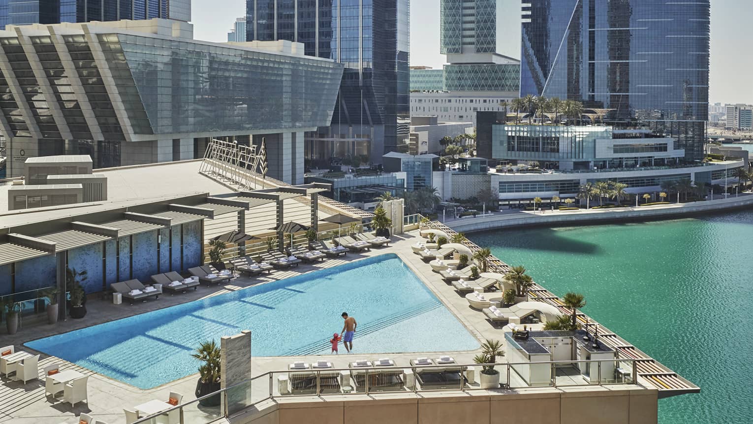 A wide shot of a man and child walking around a triangular pool overlooking the water and Abu Dhabi skyscrapers in the background. 