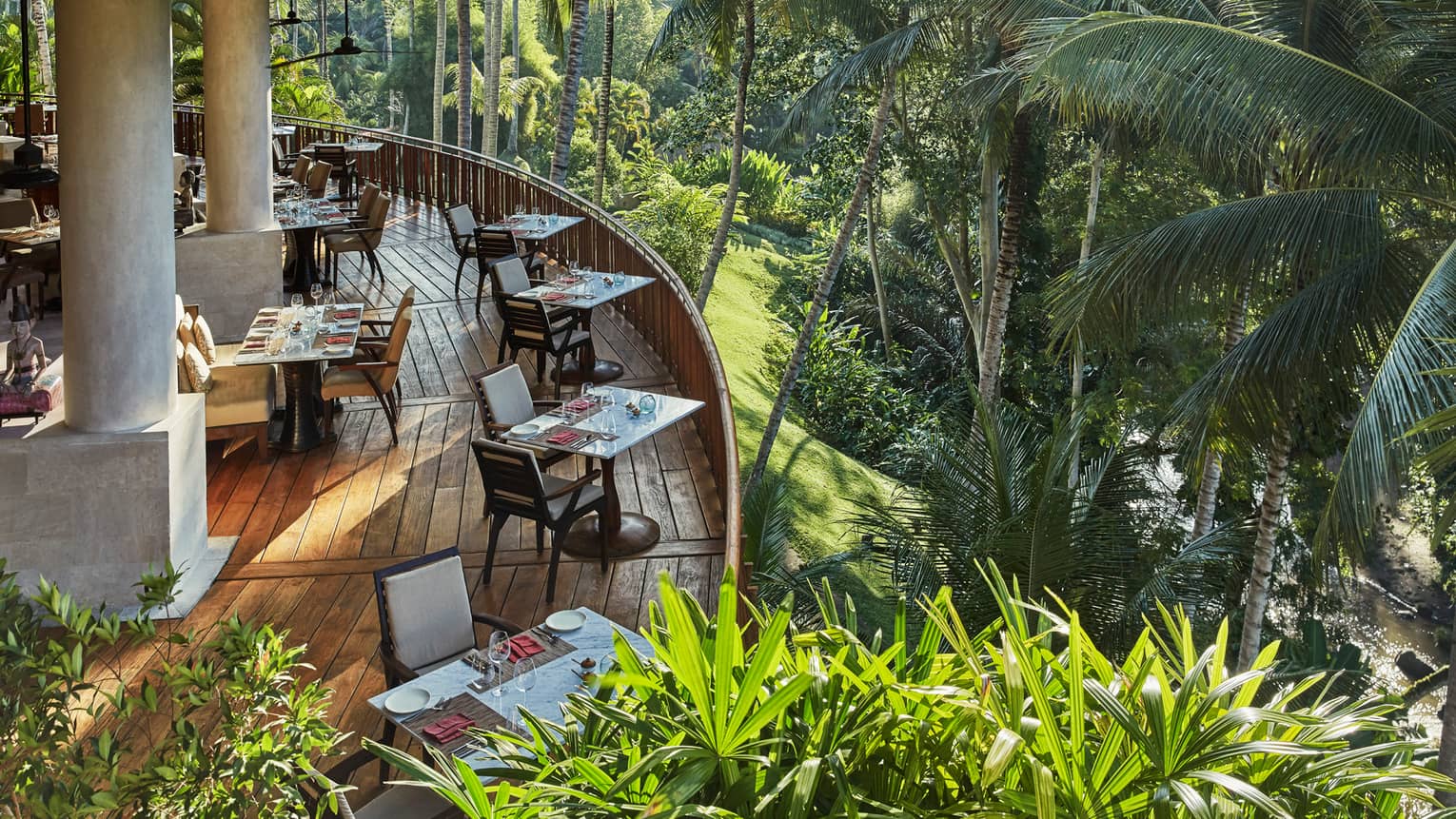 Looking down at curved wood patio of Ayung Terrace outdoor dining room, tables and chairs overlooking palm trees, forest
