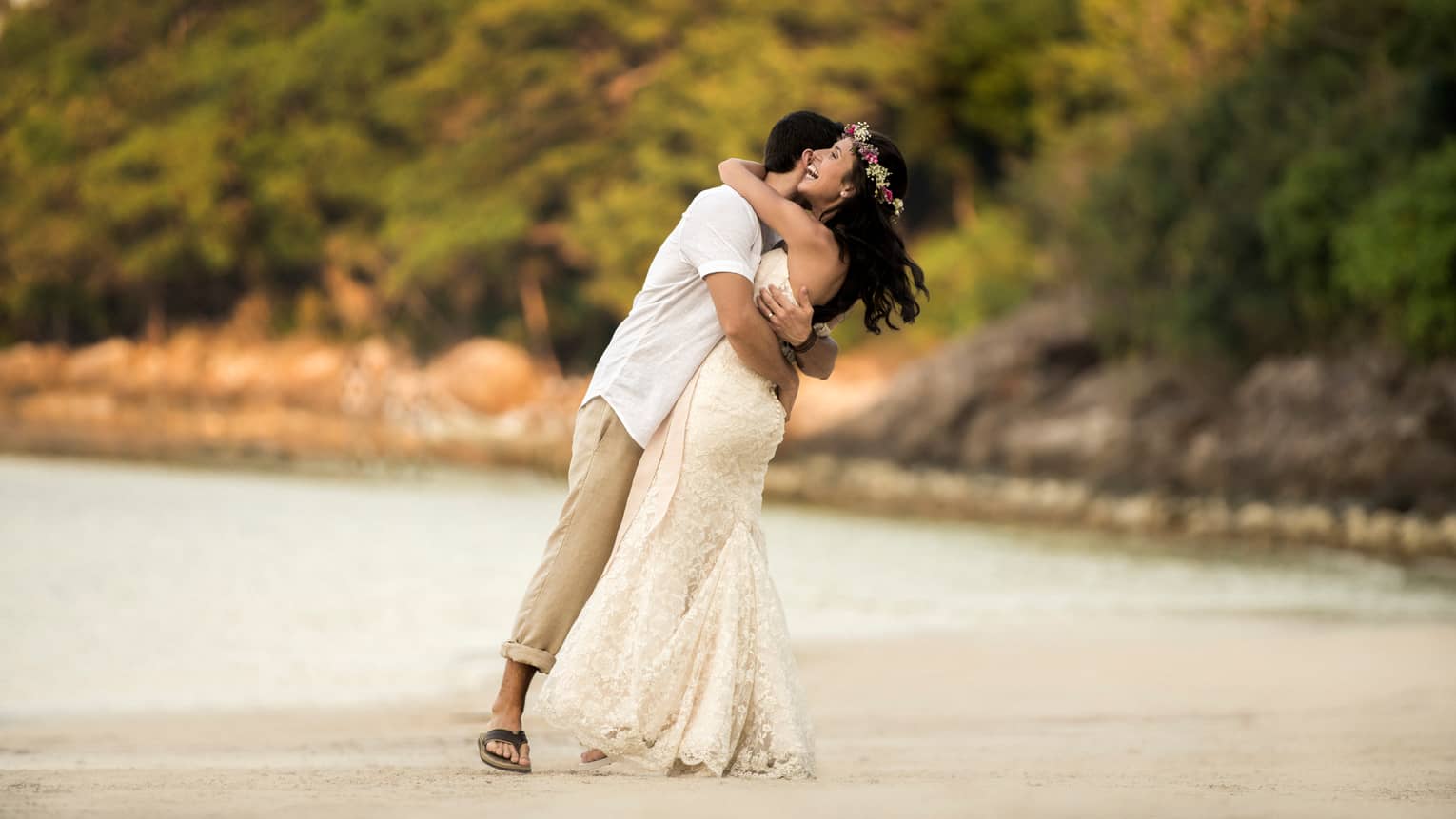Man in white shirt and khaki pants, woman in long white wedding gown and flower crown embrace on the beach