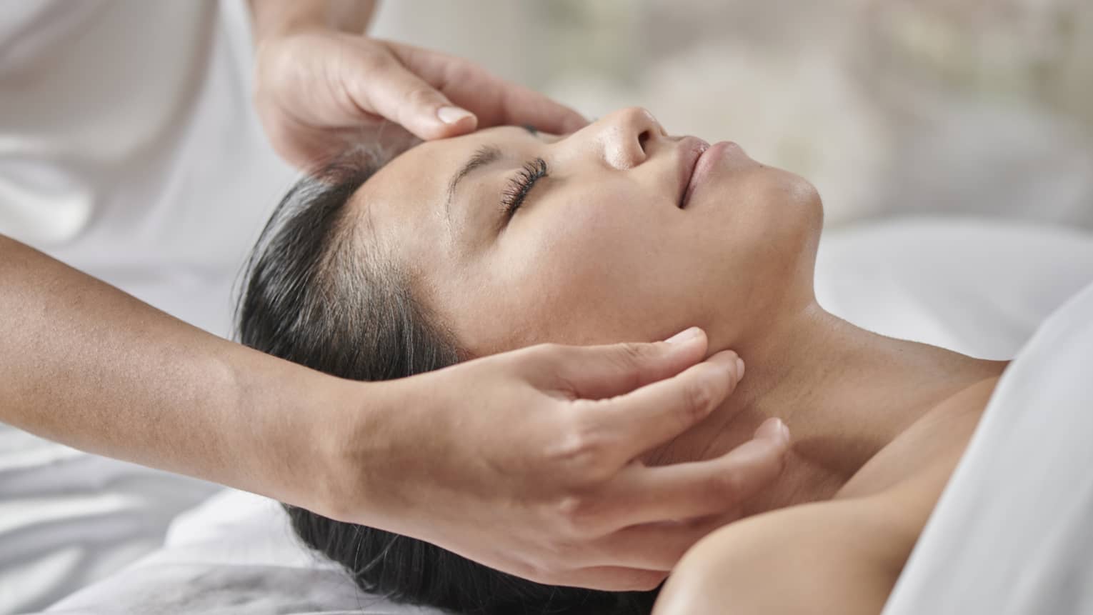 Masseuse massages woman's face as she lies face up on table under white sheet