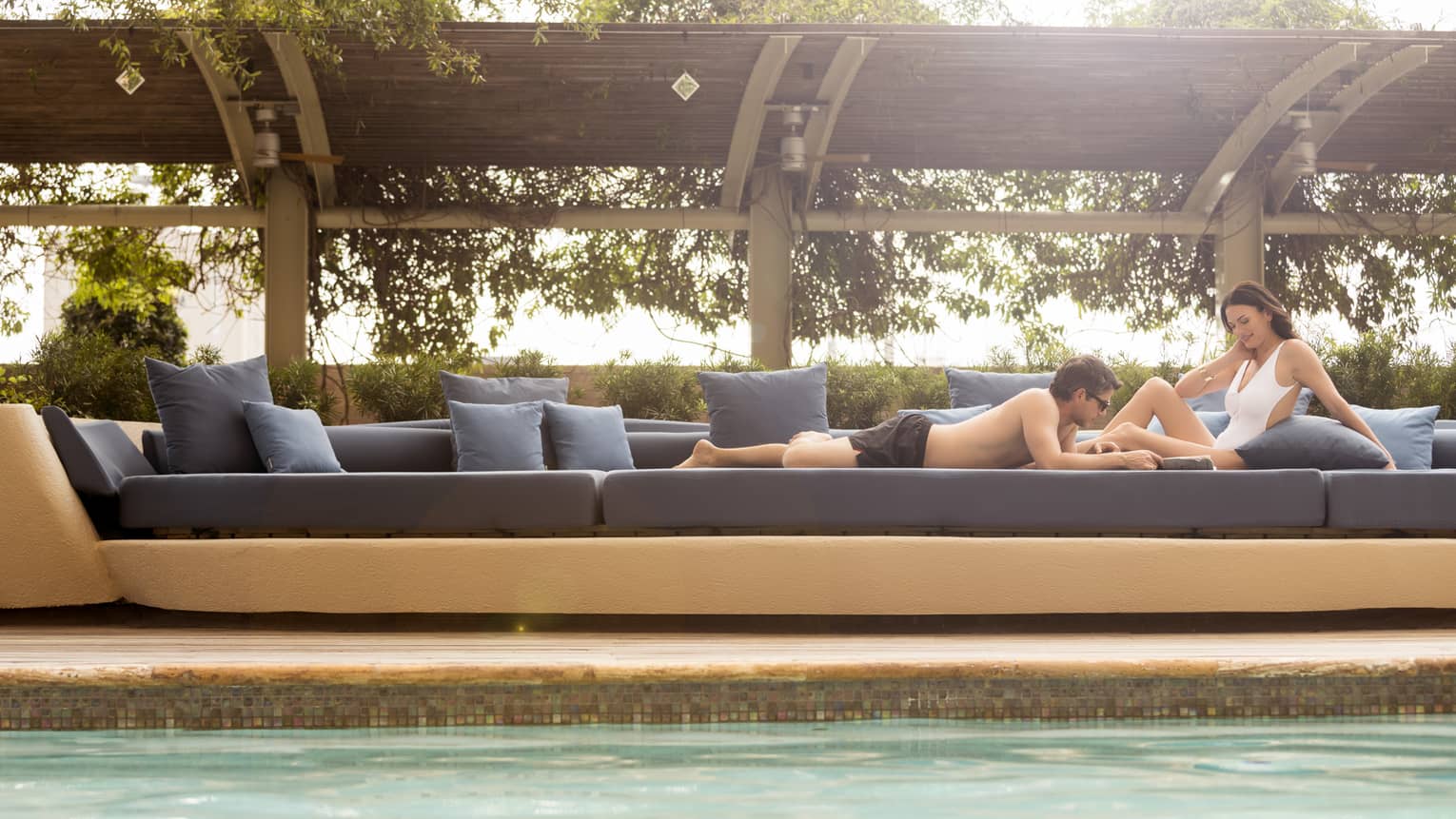 Man in swim trunks and woman in white swimsuit sunbathe on large blue patio banquette 