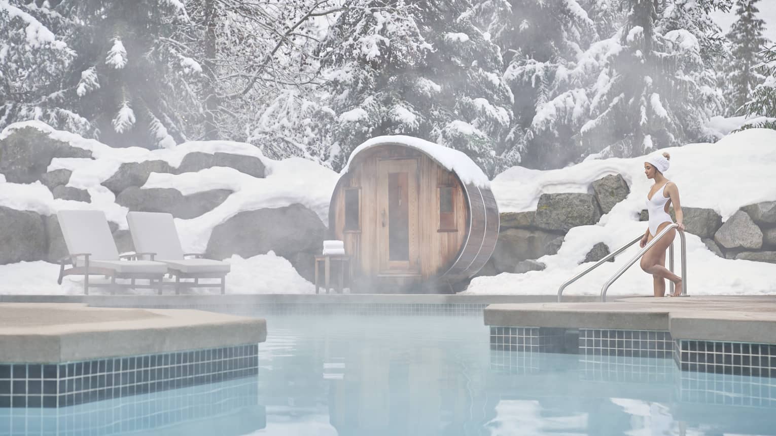 A woman in a white swimsuit enters the outdoor heated pool as steam rises around her. She is surrounded by snow-capped trees.