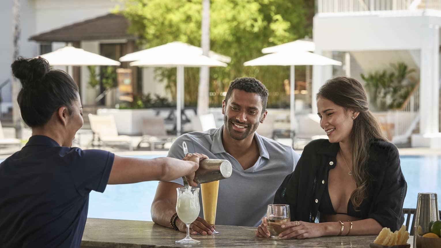 Female bartender pours a tropical drink for man and woman at poolside bar